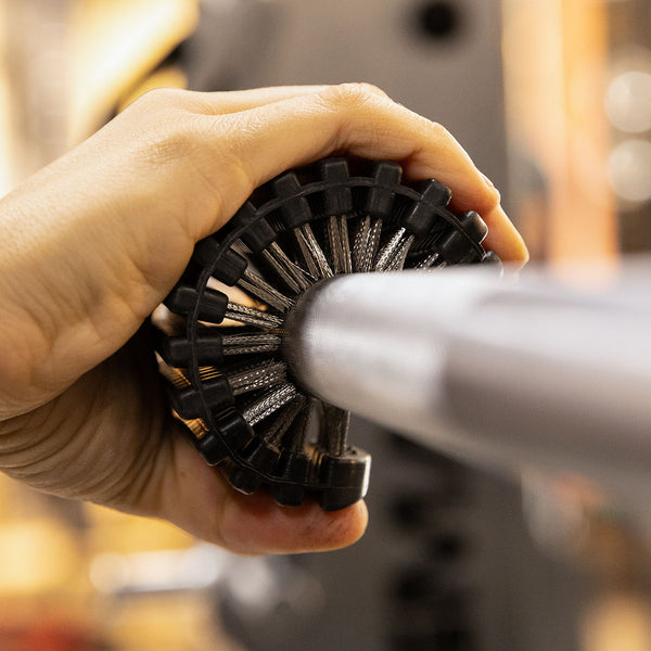 A hand uses the SMAI Barbell Cleaning Brush - Steel to scrub the knurling on a metal barbell in a gym setting.