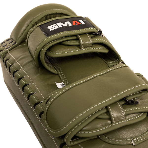 Close-up of an olive green SMAI Thai Arm Pads V2, crafted from high-grade synthetic leather with white stitching, black laces along the edges, and a velcro strap bearing the “SMAI” logo in black, white, and red. Designed with layered foam for optimal shock absorption, these pads are ideal for intense training sessions.