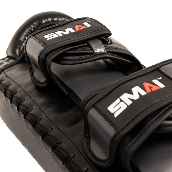 Close-up of an Essentials Muay Thai Pad (Pair) – Economy Grey by SMAI, featuring two straps labeled “SMAI” in white and red on a grey background. Crafted from durable synthetic leather, these pads are ideal for kickboxing training and showcase detailed stitching along the edges.
