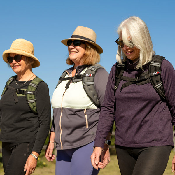Three older women, each in sunglasses, hats, backpacks, and the pink SMAI Weighted Ruck Vest, walk together outdoors on a sunny day, smiling and enjoying each other's company under a clear blue sky.