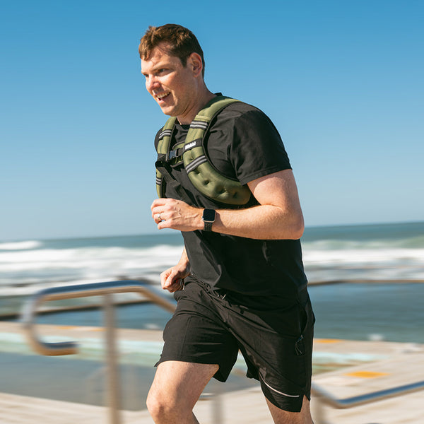 A man in black athletic clothes runs outdoors near the beach, wearing the SMAI Weighted Ruck Vest in pink and a smartwatch, with blue sky and ocean waves in the background.
