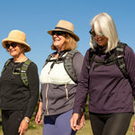Three older women, each smiling and enjoying a sunny day outdoors, ruck under a clear blue sky while wearing SMAI Weighted Vests in Olive Green.
