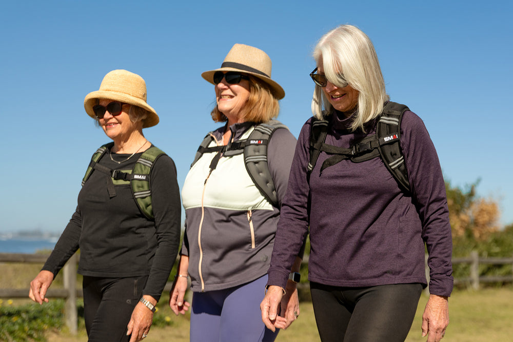 Three older women wearing hats, sunglasses, and backpacks walk outdoors on a sunny day, smiling and enjoying themselves, with a blue sky and grassy area in the background.