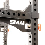 Close-up of the SMAI Folding Wall Mounted Squat Rack in black metal, featuring multiple adjustment holes and a prominent white "SMAI" logo. The rack is equipped with orange safety pins on the frame, while its robust and industrial construction makes it an excellent choice for a compact wall-mounted gym setup.