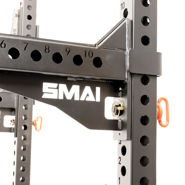 Close-up of the SMAI Folding Wall Mounted Squat Rack in black metal, featuring multiple adjustment holes and a prominent white "SMAI" logo. The rack is equipped with orange safety pins on the frame, while its robust and industrial construction makes it an excellent choice for a compact wall-mounted gym setup.