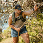 A man wearing the SMAI Weighted Vest - Black, a cap, tank top, and shorts climbs a rope outdoors in a sunlit wooded area surrounded by greenery.