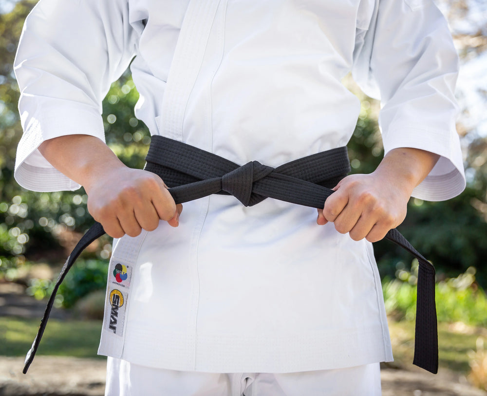 A person in a white martial arts uniform stands outdoors, holding the ends of a tied black belt at their waist.