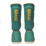 A pair of SMAI Gold Series Muay Thai Shin Guards in green with gold trim, featuring the word "SMAI" printed vertically in gold on the front of each guard. Engineered for optimal safety, these shin guards offer multi-layer EVA protection and high-density foam padding to deliver maximum comfort and impact resistance during training sessions.