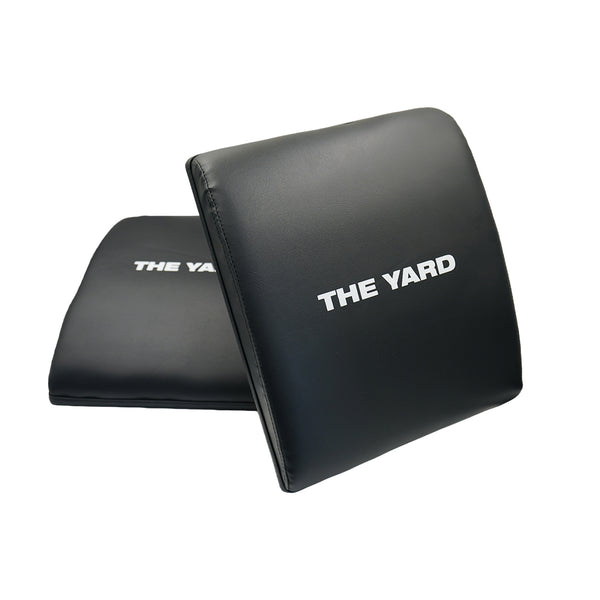 Two SMAI "The Yard - Ab Mat - Cross Training" black padded mats, each featuring "THE YARD" in white, are angled on a white background—ideal for lower back support and ab exercises in your CrossFit routine.