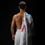 A shirtless man stands with his back to the camera against a dark background, showcasing an Olympic rings tattoo on his upper back. Draped over his left shoulder is the sleek WKF Reversible Karate Uniform – 6oz Premium Kumite Gi – Inazuma RX by SMAI, while he wears white pants. The scene exudes a sense of quiet strength and focus.