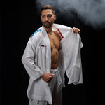 A person wearing the SMAI WKF Reversible Karate Uniform - 6oz Premium Kumite Gi - Inazuma RX stands confidently against a dark background with smoke to the side, partially opening the gi to reveal their chest. Featuring colorful patches on the shoulders, this gi embodies strength and precision.
