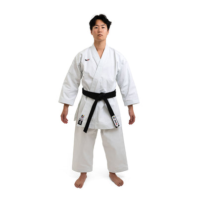 A martial artist in a white SMAI WKF Karate Uniform - Premium Kata Gi - Arashi stands barefoot on a clean backdrop, proudly displaying their black belt and patches on the left side and belt, embodying discipline and focus with a neutral expression.