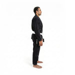 A man wears the SMAI Supreme X Brazilian Jiu Jitsu Uniform in black, featuring reinforced stitching and a white belt, standing barefoot in side profile against a plain white background.