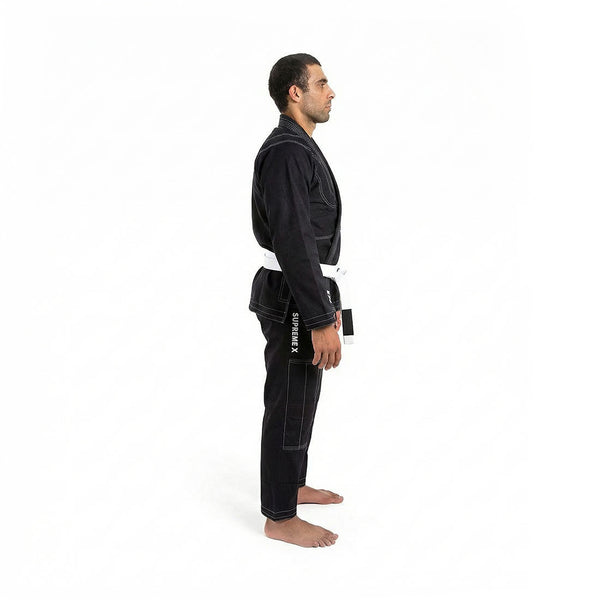 A man wears the SMAI Supreme X Brazilian Jiu Jitsu Uniform in black, featuring reinforced stitching and a white belt, standing barefoot in side profile against a plain white background.