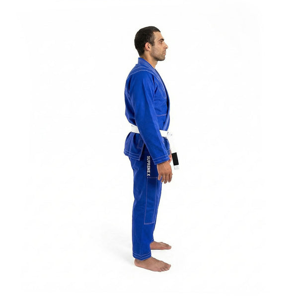 A man stands barefoot in profile, wearing the SMAI Supreme X Brazilian Jiu Jitsu Uniform in blue, made of pearl weave fabric with a white belt, against a plain white background.