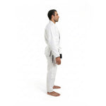 A man stands barefoot in side profile against a white background, wearing the SMAI Supreme X Brazilian Jiu Jitsu Uniform in white, made of pearl weave fabric, with black text on the pants and his arms resting by his sides.