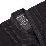 Close-up of the SMAI Supreme X Brazilian Jiu Jitsu Uniform in black, showing white stitching, a pearl weave collar, and an inside label: "SUPREME X PREMIUM BJJ A3, 65% cotton, 35% polyester," care symbols, and the SMAI brand name.