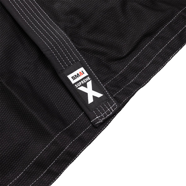 Close-up of the SMAI Supreme X Brazilian Jiu Jitsu Uniform in black, featuring white stitching and durable pearl weave fabric. The "SMAI SUPREME X" tag appears in black, white, and red, highlighting the uniform’s textured details.