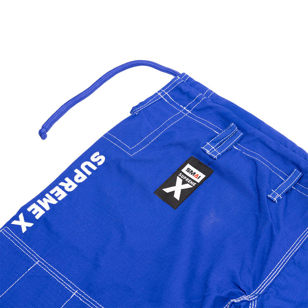 Close-up of SMAI Supreme X Brazilian Jiu Jitsu Uniform blue pearl weave pants with white stitching, "SUPREMEX" in white text, a black and red SMAI patch near a large "X," and a blue drawstring at the waistband.