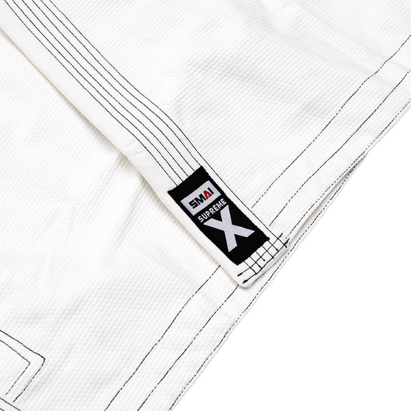 Close-up of the SMAI Supreme X Brazilian Jiu Jitsu Uniform in white, featuring black stitching, a "SMAI Supreme X" lapel patch, textured pearl weave fabric, and reinforced seams for durability.