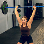 A woman in a black tank top and shorts lifts a 20kg blue SMAI barbell overhead in a gym, focusing intently on her workout.