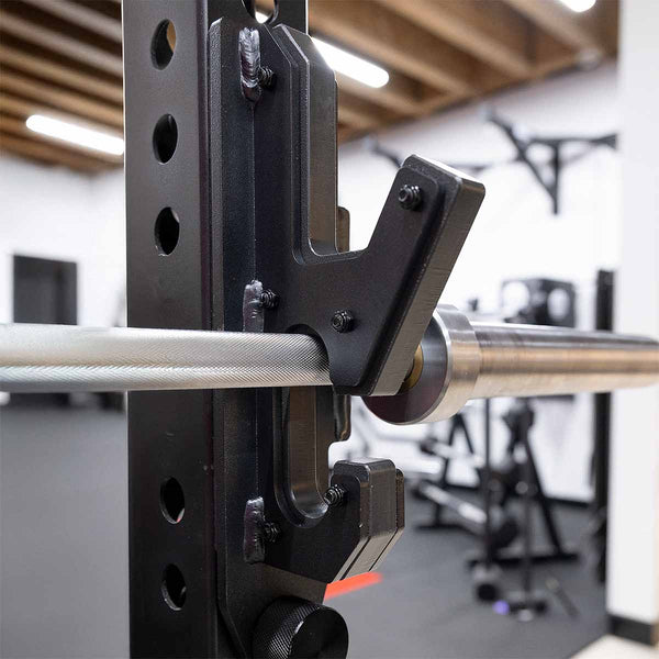 Close-up of the SMAI 2-in-1 Isometric Cup / J-Hook Utility attached to a Vanta Series Power Rack in a gym, with other workout equipment and wooden ceiling beams visible in the blurred background.