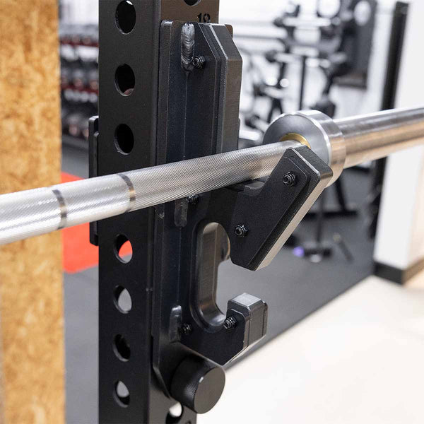 Close-up of the SMAI 2-in-1 Isometric Cup / J-Hook Utility for Vanta Series Power Racks attached to a squat rack, with gym equipment and weights visible in the blurred background.