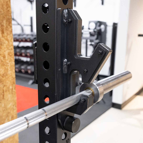 A close-up of the SMAI 2-in-1 Isometric Cup / J-Hook Utility, attached to a Vanta Series Power Rack in a gym, with weight racks and other equipment visible in the background.