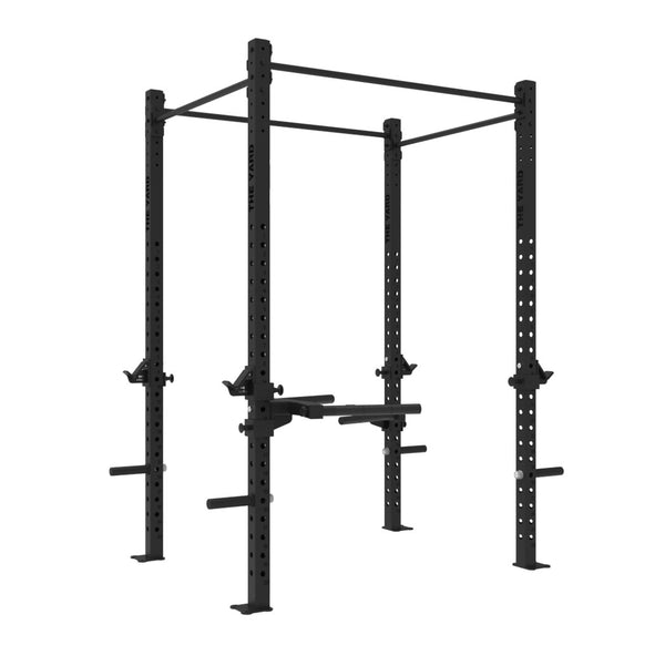 The SMAI The Yard - Rig - 2 Cell is a black metal power rack for weightlifting, designed with multiple holes for adjustable attachments and equipped with a pull-up bar at the top. It features integrated weight plate storage and safety bars, as well as convenient weight plate holders. This freestanding squat rig can also be fitted with optional dip stations, all presented against a white background.