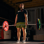A man in a black T-shirt and green shorts performs a deadlift with SMAI Calibrated Weight Plates - 10kg (Pair) on a loaded barbell in a gym, standing on a mat with other gym equipment visible in the background.