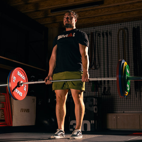 A man in a black T-shirt and green shorts lifts a barbell loaded with SMAI Calibrated Weight Plates - 15kg (Pair) in a gym featuring a wooden ceiling, black flooring, and various workout equipment.