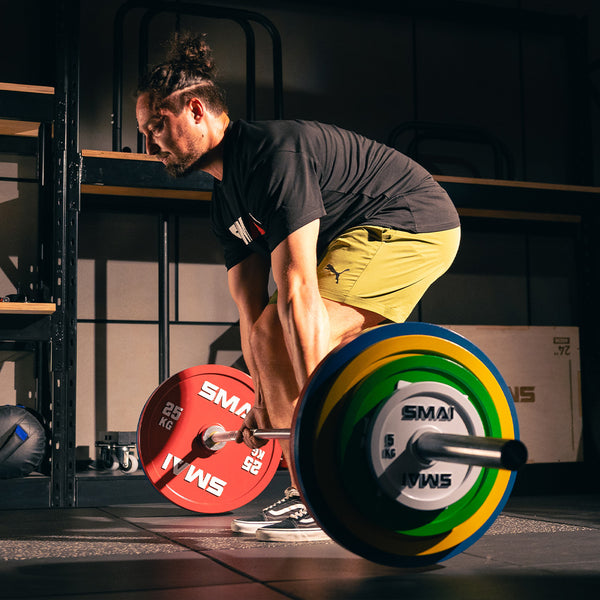 A man in a black shirt and yellow shorts prepares to deadlift a barbell loaded with SMAI Calibrated Weight Plates - 10kg (Pair) in a gym.