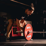 Wearing athletic gear, a person grips a barbell loaded with SMAI Calibrated Weight Plates - 25kg (Pair) in a dimly lit gym, focused and ready to lift.