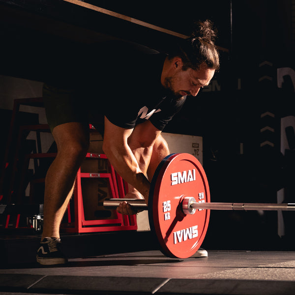 Wearing athletic gear, a person grips a barbell loaded with SMAI Calibrated Weight Plates - 25kg (Pair) in a dimly lit gym, focused and ready to lift.