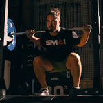 A bearded man with tied-back hair squats in a gym, wearing a black SMAI t-shirt and shorts, with a barbell loaded with SMAI Calibrated Weight Plates - 20kg (Pair) across his shoulders.