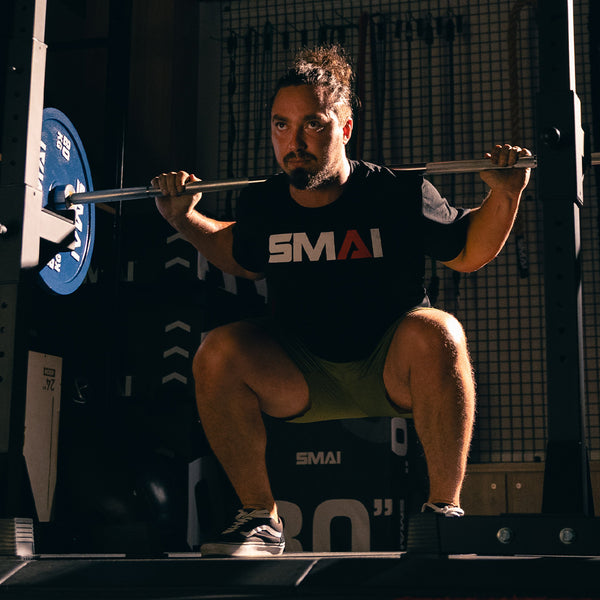 A bearded man with tied-back hair squats in a gym, wearing a black SMAI t-shirt and shorts, with a barbell loaded with SMAI Calibrated Weight Plates - 20kg (Pair) across his shoulders.