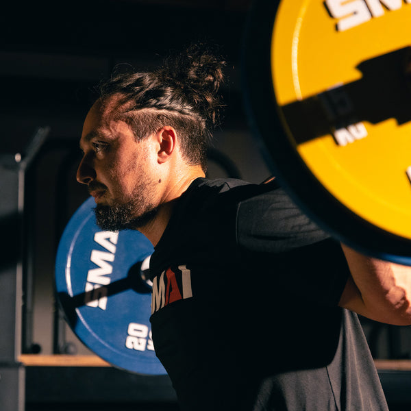 A man with a braided hairstyle and beard lifts a barbell loaded with SMAI Calibrated Weight Plates - 20kg (Pair) in a gym under dramatic lighting.