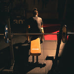A person sits on the SMAI Commercial Adjustable Bench - Tan, facing away, with a barbell racked in front of them. Dramatic shadows accentuate the weightlifting area in the dimly lit gym.