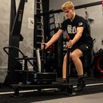 A young man with short blond hair, in a black "Team Reflex" outfit, pushes the SMAI Prowler Sled 2.0 across the gym floor, with fitness equipment and punching bags visible in the background.
