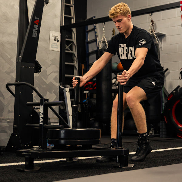 A young man with short blond hair, in a black "Team Reflex" outfit, pushes the SMAI Prowler Sled 2.0 across the gym floor, with fitness equipment and punching bags visible in the background.