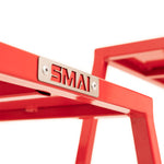 Close-up of a red metal frame from the SMAI Steel Plyo Box Set - 4 Pack Stackable, featuring a silver "SMAI" plate fastened with two screws; another red frame appears blurred in the background.