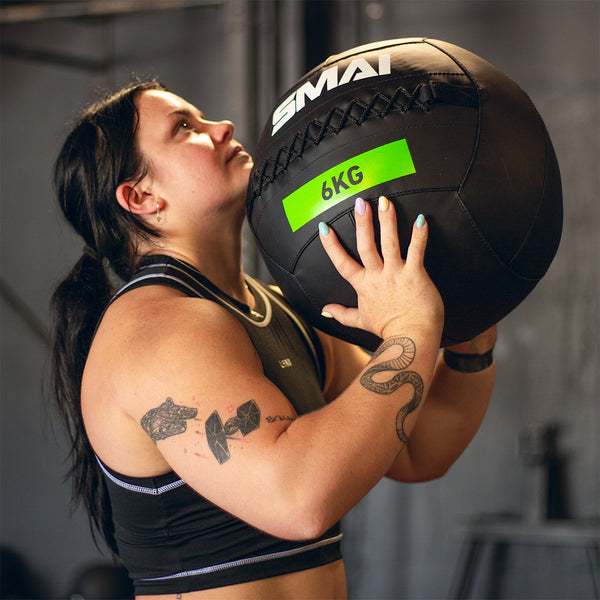Wearing athletic gear, a person with visible arm tattoos holds a black 6 kg SMAI Wall Ball and looks up, preparing to exercise in the gym.