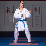 A person is wearing the WKF Reversible Karate Uniform - 6oz Premium Kumite Gi - Inazuma RX by SMAI, with a blue belt. They stand in a fighting stance on a dojo mat, with motivational posters visible on the wall behind them.