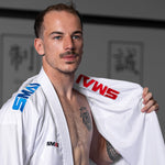 A man stands in a dojo wearing the SMAI WKF Reversible Karate Uniform - 6oz Premium Kumite Gi Inazuma RX, featuring red and blue lettering. He holds open the martial arts gi like a reversible jacket, showcasing his chest tattoo, while blurred wall art and calligraphy are visible in the background.