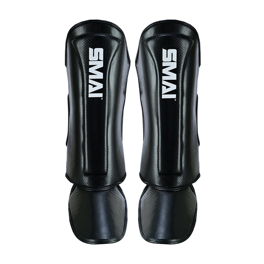 A pair of SMAI Essentials Muay Thai Shin Guards for kids in black, with foot protection and the white "SMAI" logo on the front, shown side by side on a plain white background—ideal for young athletes.