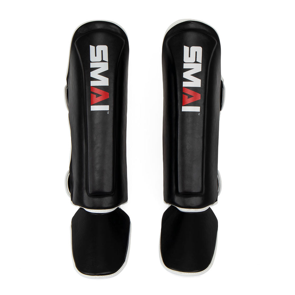 A pair of black Essentials Muay Thai Shin Guards for kids by SMAI, each featuring a white and red “SMAI” logo, with attached foot protectors—shown on a white background and designed to provide ideal martial arts protection for children.