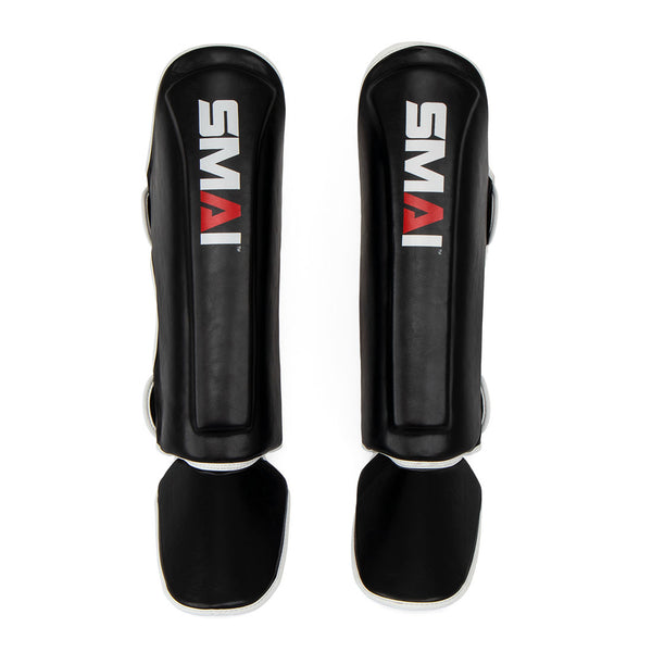 A pair of black Essentials Muay Thai Shin Guards for kids by SMAI, each featuring a white and red “SMAI” logo, with attached foot protectors—shown on a white background and designed to provide ideal martial arts protection for children.