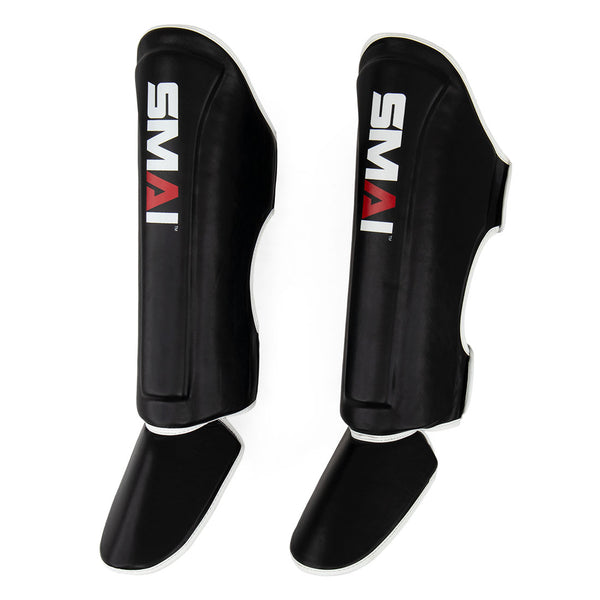 Introducing the Essentials Muay Thai Shin Guards - Kids by SMAI—a pair of black shin instep guards with attached foot protection, featuring the distinctive white and red “SMAI” logo on the front—ideal for protecting young martial artists.