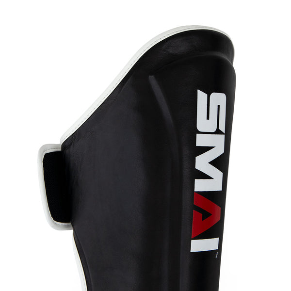 A close-up of a black Essentials Muay Thai Shin Guard - Kids by SMAI, featuring white trim and the brand name “SMAI” printed in bold white and red lettering on the front, designed for versatile protection as both shin instep guards and kids shin guards.
