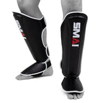 A person stands barefoot against a white background wearing black SMAI Essentials Muay Thai Shin Guards - Kids, featuring white trim and designed to cover the shins and instep for martial arts.